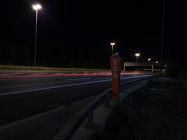 A nighttime highway scene with bright streetlights illuminating the road. An SOS emergency call box stands prominently on the roadside. Light trails from passing vehicles create streaks of color on the asphalt.