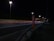 A nighttime highway scene with bright streetlights illuminating the road. An SOS emergency call box stands prominently on the roadside. Light trails from passing vehicles create streaks of color on the asphalt.