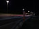 A nighttime highway scene with bright streetlights illuminating the road. An SOS emergency call box stands prominently on the roadside. Light trails from passing vehicles create streaks of color on the asphalt.