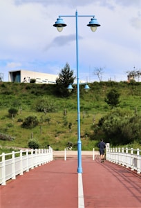 A person is jogging along a red-colored path bordered by white railings, leading towards a green, grassy hill. A blue lamp post stands prominently in the center of the path. In the background, a modern white building and a cyclist can be seen on top of the hill.