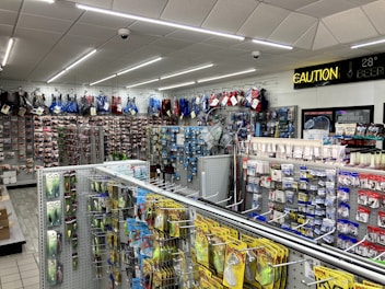 A well-lit store interior filled with fishing and outdoor equipment. The shelves are stocked with various lures, fishing lines, and other accessories. A range of colorful life vests is displayed on the back wall. A caution sign and a digital temperature display are mounted near the ceiling.