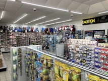 A well-lit store interior filled with fishing and outdoor equipment. The shelves are stocked with various lures, fishing lines, and other accessories. A range of colorful life vests is displayed on the back wall. A caution sign and a digital temperature display are mounted near the ceiling.