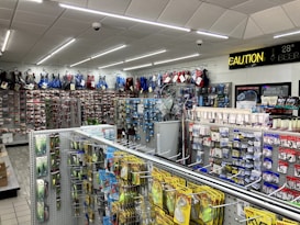 A well-lit store interior filled with fishing and outdoor equipment. The shelves are stocked with various lures, fishing lines, and other accessories. A range of colorful life vests is displayed on the back wall. A caution sign and a digital temperature display are mounted near the ceiling.