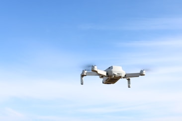 A white drone is flying in the sky, capturing a clear image of its four rotors. The background features a light blue sky with a few wispy clouds.