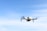 A white drone is flying in the sky, capturing a clear image of its four rotors. The background features a light blue sky with a few wispy clouds.