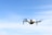 A white drone is flying in the sky, capturing a clear image of its four rotors. The background features a light blue sky with a few wispy clouds.