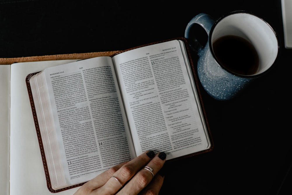 Person liest Buch neben weißer Keramiktasse mit Kaffee