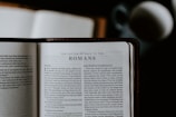 An open Bible lies on a flat surface, displaying the text of 'The Letter of Paul to the Romans.' A blurred cup or mug is visible in the background, suggesting a casual reading environment.