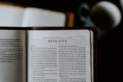 An open Bible lies on a flat surface, displaying the text of 'The Letter of Paul to the Romans.' A blurred cup or mug is visible in the background, suggesting a casual reading environment.