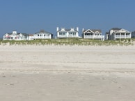 Several beachfront houses stand in a row behind a stretch of sandy beach and dune grass. The sky is clear blue, and the scene is calm and serene.