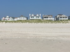 Several beachfront houses stand in a row behind a stretch of sandy beach and dune grass. The sky is clear blue, and the scene is calm and serene.