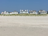 Several beachfront houses stand in a row behind a stretch of sandy beach and dune grass. The sky is clear blue, and the scene is calm and serene.