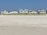 Several beachfront houses stand in a row behind a stretch of sandy beach and dune grass. The sky is clear blue, and the scene is calm and serene.
