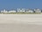 Several beachfront houses stand in a row behind a stretch of sandy beach and dune grass. The sky is clear blue, and the scene is calm and serene.