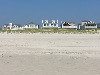 Several beachfront houses stand in a row behind a stretch of sandy beach and dune grass. The sky is clear blue, and the scene is calm and serene.