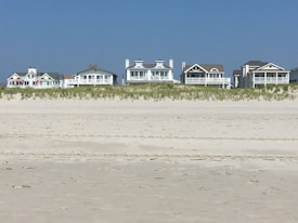 Several beachfront houses stand in a row behind a stretch of sandy beach and dune grass. The sky is clear blue, and the scene is calm and serene.