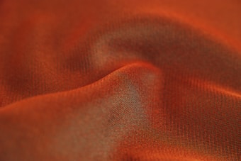 A close-up of a textured fabric with a wavy pattern, predominantly featuring a warm orange hue. The fabric appears soft and slightly translucent, with light and shadow creating a sense of depth and movement.