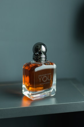 A glass bottle of perfume with a dark metallic cap, resting on a smooth, gray surface. The bottle contains an amber-colored liquid and features the text 'Stronger With You Intensely' on its front.
