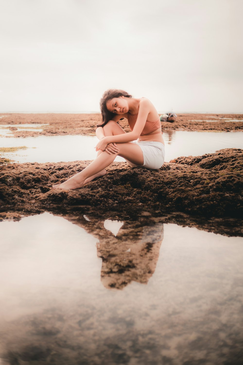 Mujer en camiseta blanca sin mangas sentada en roca marrón cerca del cuerpo de agua durante el día