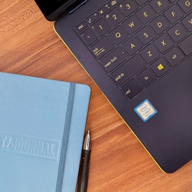 A close-up view of a laptop keyboard with yellow symbols alongside a light blue journal and a pen, all placed on a wooden surface. The laptop has a visible Intel Core i7 sticker.