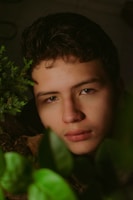 A young person is surrounded by lush green foliage. The light is soft, highlighting the serene and contemplative expression on their face. The setting and close proximity to the plants suggest a connection with nature.