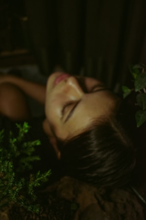 A person with closed eyes surrounded by lush green leaves and dark shadows, creating a peaceful and contemplative atmosphere.