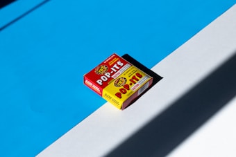 A red and yellow box of Pop-Its, novelty firecrackers, is placed on a surface divided by a shadow. The background features a stark contrast of bright blue and white surfaces, enhancing the vivid colors of the box. The lighting creates strong shadows, adding depth to the image.