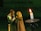 A couple in medieval attire stands close together, appearing engaged in an intimate moment. The man wears chainmail and a breastplate, while the woman draped in a cloak interacts softly with him. A lit candle on a holder is prominently placed in the foreground, casting a warm light.