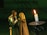 A couple in medieval attire stands close together, appearing engaged in an intimate moment. The man wears chainmail and a breastplate, while the woman draped in a cloak interacts softly with him. A lit candle on a holder is prominently placed in the foreground, casting a warm light.