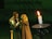 A couple in medieval attire stands close together, appearing engaged in an intimate moment. The man wears chainmail and a breastplate, while the woman draped in a cloak interacts softly with him. A lit candle on a holder is prominently placed in the foreground, casting a warm light.