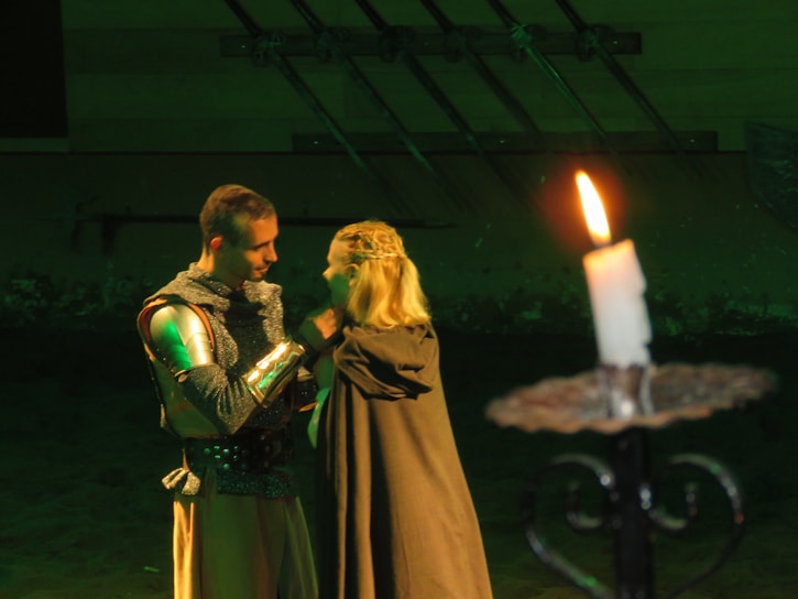 A couple in medieval attire stands close together, appearing engaged in an intimate moment. The man wears chainmail and a breastplate, while the woman draped in a cloak interacts softly with him. A lit candle on a holder is prominently placed in the foreground, casting a warm light.