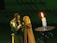 A couple in medieval attire stands close together, appearing engaged in an intimate moment. The man wears chainmail and a breastplate, while the woman draped in a cloak interacts softly with him. A lit candle on a holder is prominently placed in the foreground, casting a warm light.