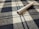 A cigarette lies on a fabric surface with a checkered pattern, featuring blue and white colors. The textile texture is clearly visible, and the cigarette appears unused with the tobacco showing at one end.