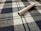 A cigarette lies on a fabric surface with a checkered pattern, featuring blue and white colors. The textile texture is clearly visible, and the cigarette appears unused with the tobacco showing at one end.