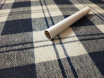 A cigarette lies on a fabric surface with a checkered pattern, featuring blue and white colors. The textile texture is clearly visible, and the cigarette appears unused with the tobacco showing at one end.