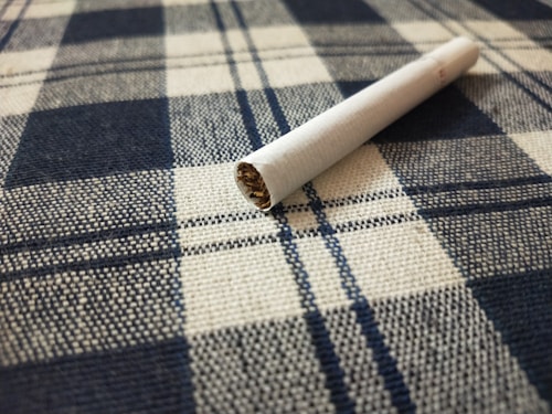 A cigarette lies on a fabric surface with a checkered pattern, featuring blue and white colors. The textile texture is clearly visible, and the cigarette appears unused with the tobacco showing at one end.