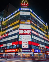 A brightly illuminated multi-story building features numerous colorful signs and advertisements in various languages, including large McDonald's logos and signage for hotels and other businesses.