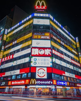 A brightly illuminated multi-story building features numerous colorful signs and advertisements in various languages, including large McDonald's logos and signage for hotels and other businesses.