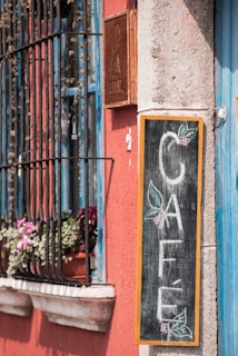 A colorful storefront with a chalkboard sign displaying the word 'CAFE' alongside artistic leaf and berry drawings. The building features a vibrant red facade, a blue door, and a flower box with pink and purple flowers. Metal bars cover the window.