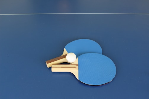 Two blue ping pong paddles and a white ping pong ball on a blue table with a visible net line in the background.