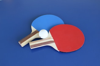 Two table tennis paddles, one with a blue rubber face and the other with a red rubber face, are placed on a blue surface. A white ping pong ball is positioned between the paddles.