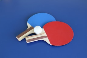 Two table tennis paddles, one with a blue rubber face and the other with a red rubber face, are placed on a blue surface. A white ping pong ball is positioned between the paddles.