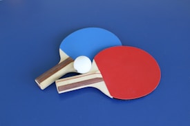 Two table tennis paddles, one with a blue rubber face and the other with a red rubber face, are placed on a blue surface. A white ping pong ball is positioned between the paddles.
