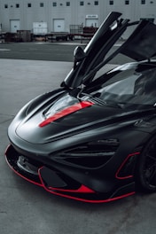 A sleek, high-performance sports car with a black matte finish and red accents is parked in an industrial area. The car's doors are open upwards, showcasing its modern and aerodynamic design. The background contains industrial buildings and storage equipment.