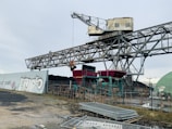 An industrial scene featuring a large steel structure resembling a conveyor belt system, possibly used for material handling. Graffiti is visible on a nearby concrete wall, and a construction-like environment is present with metal fences and piles of materials in the foreground. The sky is overcast, creating a muted atmosphere.