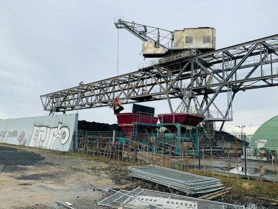 An industrial scene featuring a large steel structure resembling a conveyor belt system, possibly used for material handling. Graffiti is visible on a nearby concrete wall, and a construction-like environment is present with metal fences and piles of materials in the foreground. The sky is overcast, creating a muted atmosphere.
