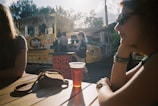 A food truck decorated with vibrant graphics is parked outdoors on a sunny day. Three people are nearby, with one leaning on a table in the foreground, wearing sunglasses. On the table, there is a drink in a plastic cup and a small bag placed beside it. Another person is walking past the truck, which has signage advertising different sauces. Trees and a partly cloudy sky are visible in the background.
