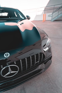 A close-up view of the front section of a sleek black Mercedes-Benz car with the emblem prominently displayed. The car is parked in an outdoor setting, and there is a reflection of the surroundings on its shiny surface.
