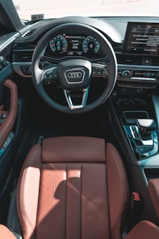 A luxurious car interior featuring a leather seat and a detailed steering wheel with the Audi logo prominently displayed. Dashboard displays include modern digital interfaces and controls. The setting is well-organized and technologically advanced.