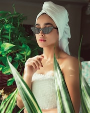 A person wearing a towel turban and fashionable sunglasses is surrounded by lush green plants. The setting is serene, with soft lighting enhancing their contemplative expression. The person has a spa-like appearance, adding a sense of relaxation to the atmosphere.